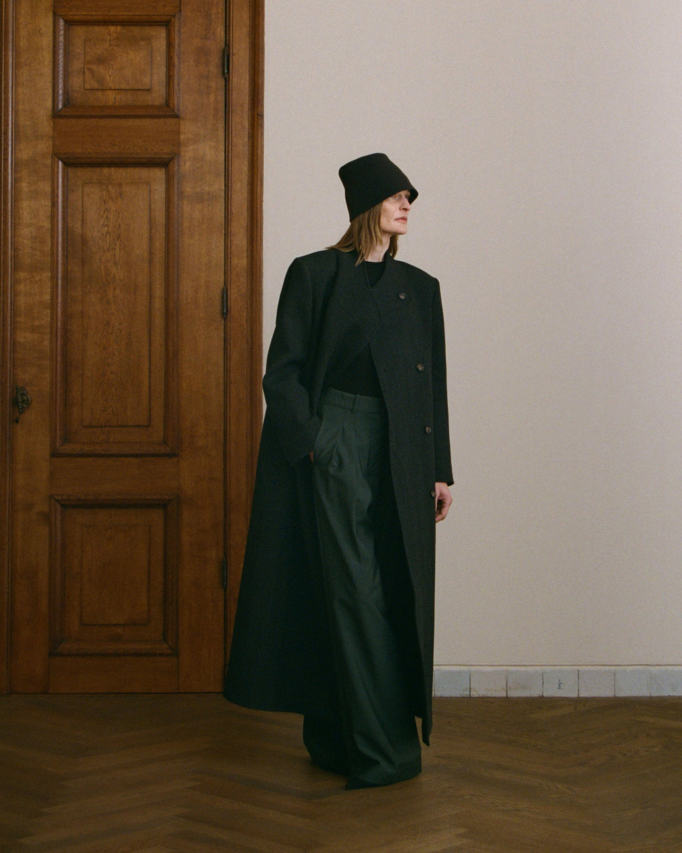 Woman wearing a long dark coat, dark green trousers and a black hat standing in a room with wooden floor and door.