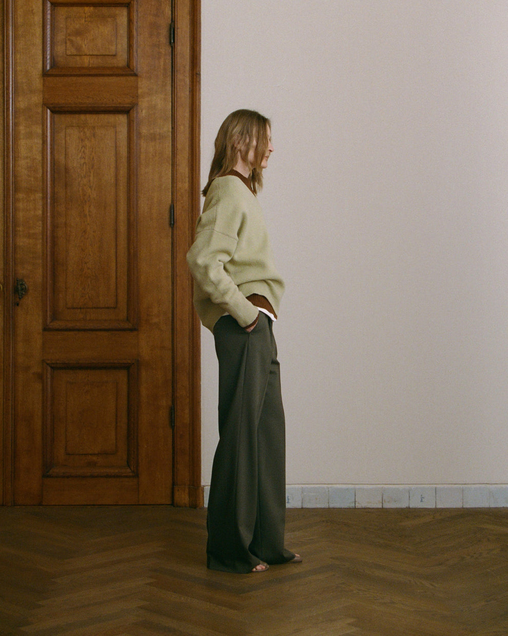 Woman in a light green sweater and dark green pants standing in a room with wooden flooring and a wooden door.