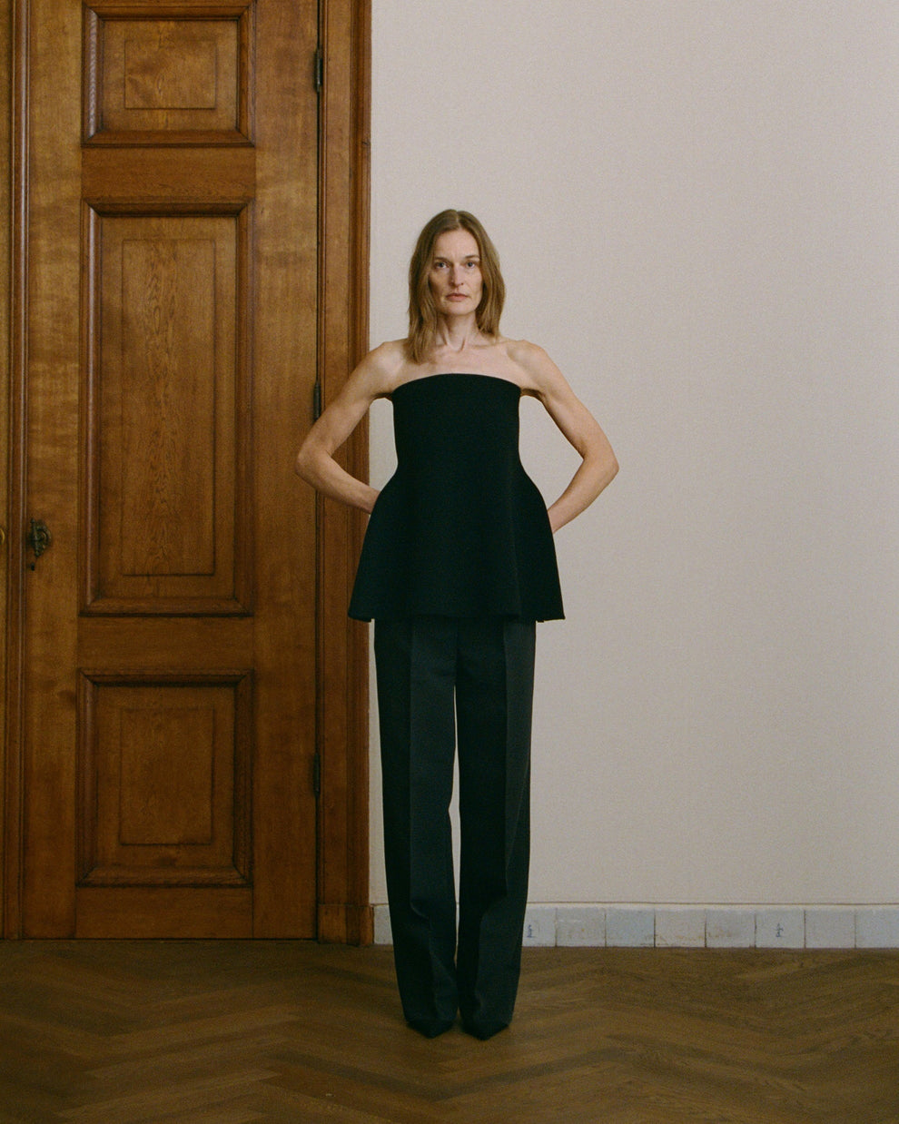 Woman wearing a black strapless top and high-waisted black pants standing in a room with wooden paneling.