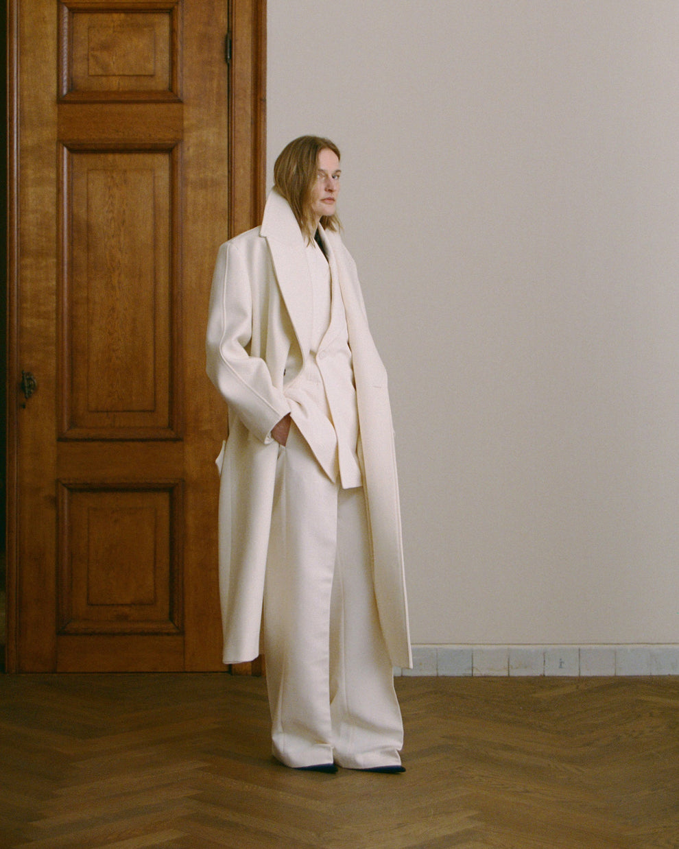 Woman wearing a long cream coat, cream blazer and cream pants standing in a room with wooden flooring and a wooden door.