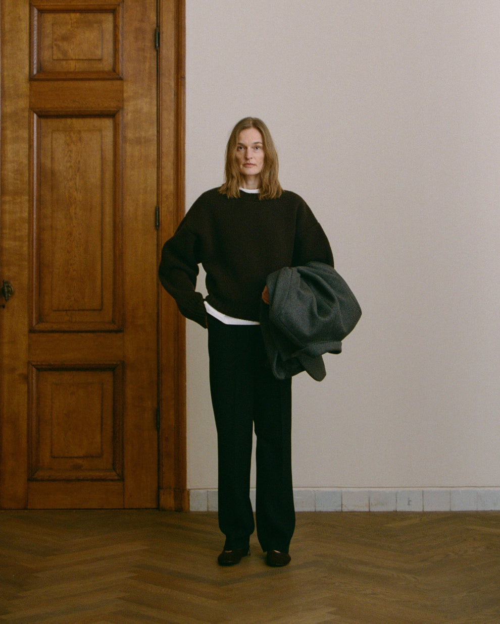 Woman wearing a dark brown thick jumper holding a grey coat in a room with wooden door and light-colored wall