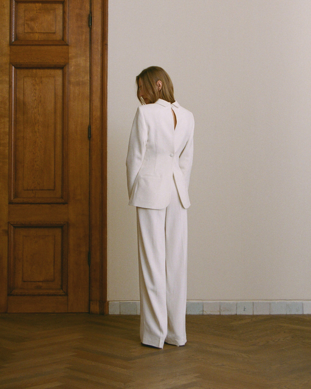 Woman wearing a white backwards blazer and matching white trousers standing in a room with wooden floors and paneling.