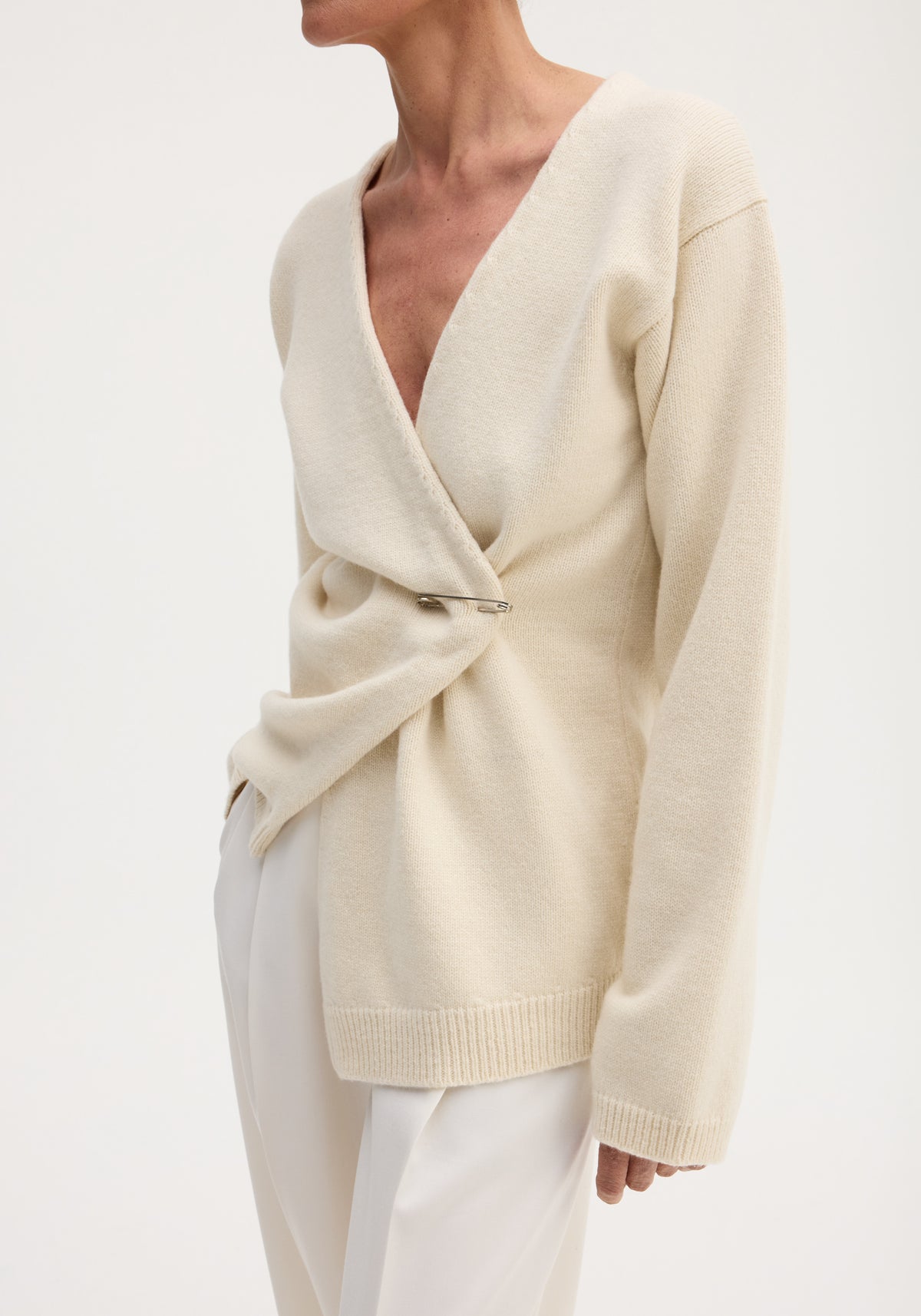 Off-shoulder wrap cardigan in cream, featuring a deep V-neck, safety-pin closure, long sleeves, worn with off-white trousers for a minimal, monochromatic look.
