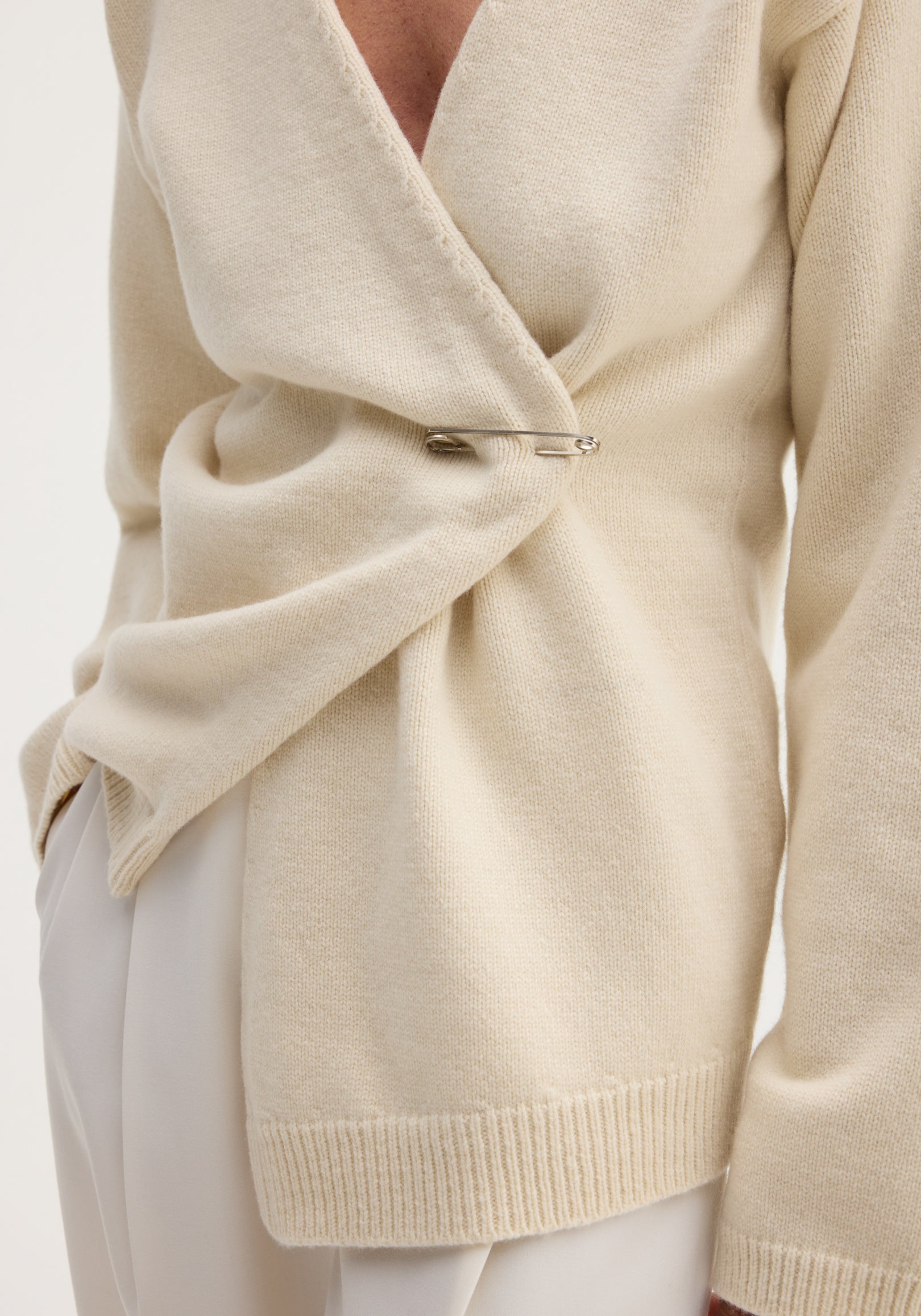 Off-shoulder wrap cardigan in cream, draped with a silver safety pin closure, crafted from a soft wool-cashmere blend, paired with off-white trousers.