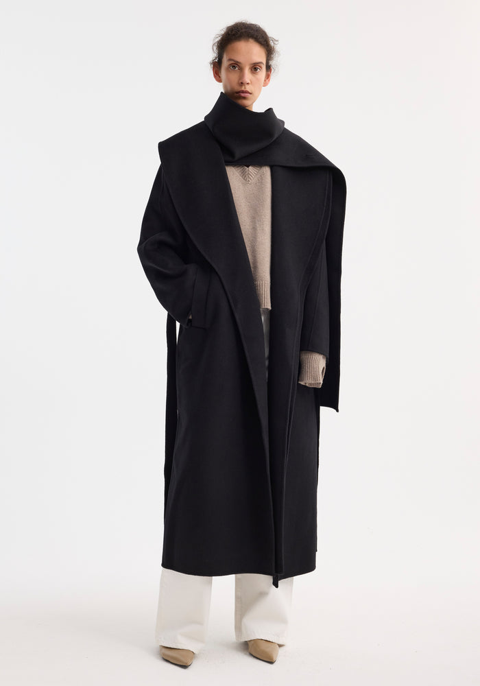 Model wearing Signature double-faced scarf coat with belt, showcasing its oversized collar, side pockets, and wrap belt, layered over a V-neck sweater and white trousers.
