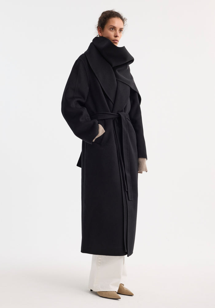 Signature double-faced scarf coat with belt in black, worn by a woman. Features oversized collar, belted waist, side pockets; styled with white trousers, nude heels.