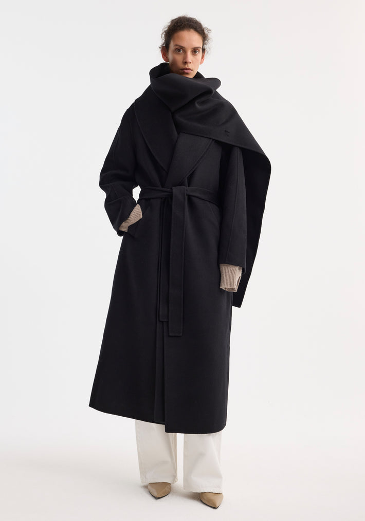 Model wearing the Signature double-faced scarf coat with belt, showcasing its sculptural scarf, wrap style, and taupe fingerless gloves, paired with white trousers and beige heels.