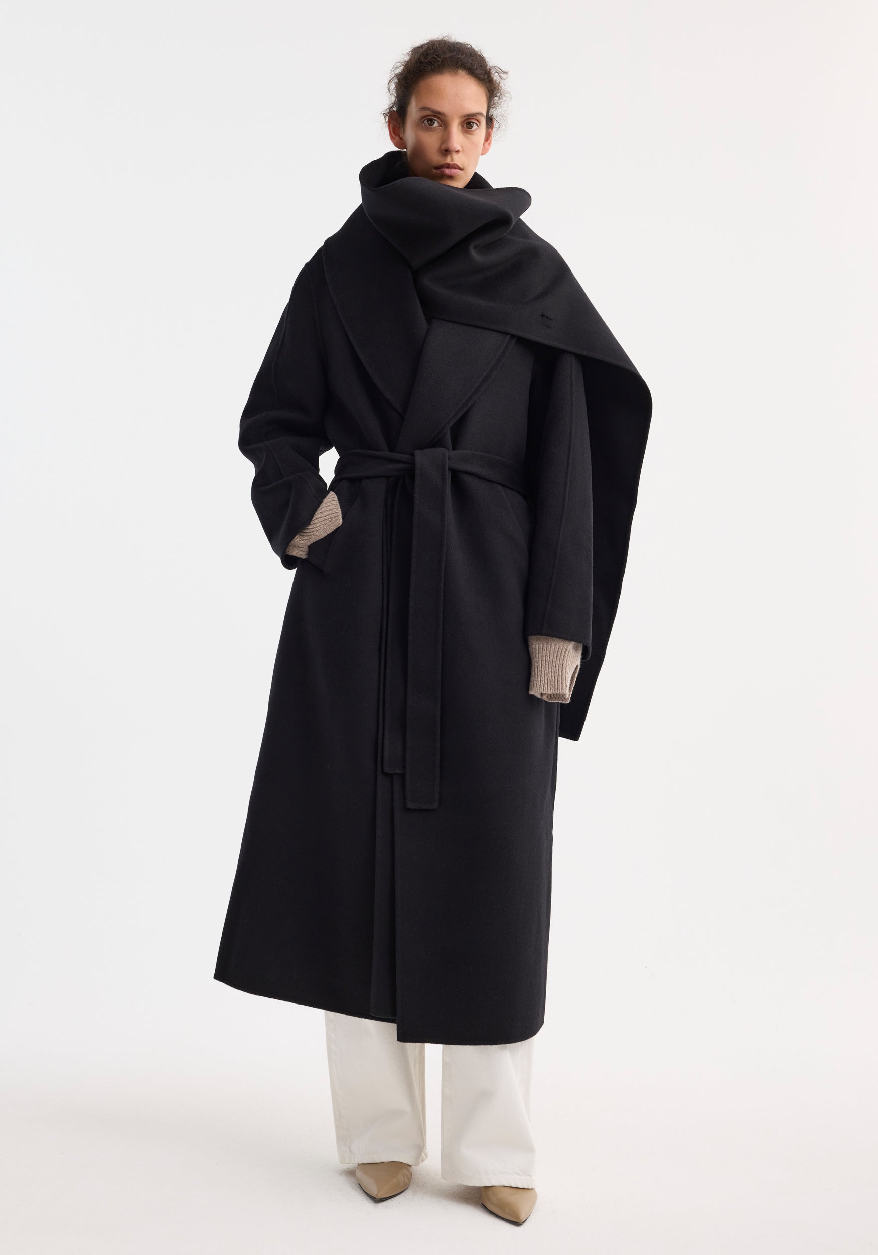 Woman wearing a black Signature double-faced scarf coat with belt | black, styled with wide scarf detail, beige gloves, white pants, and beige shoes against a plain background.