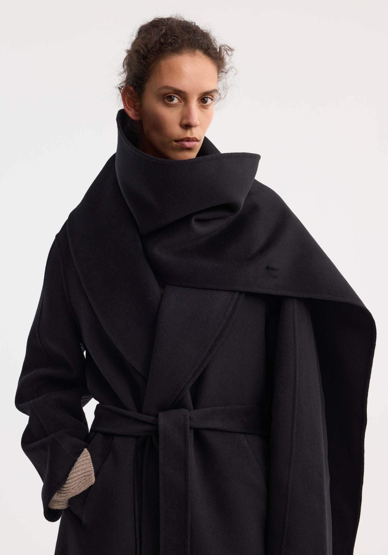 Woman wearing a black Signature double-faced scarf coat with belt | black, featuring an oversized scarf collar and tied waist belt, against a plain light background.