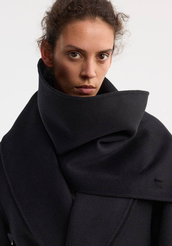Signature double-faced scarf coat in black, showcasing its voluminous wool fabric draping elegantly around the model’s shoulders, with visible wrap belt and side pockets.