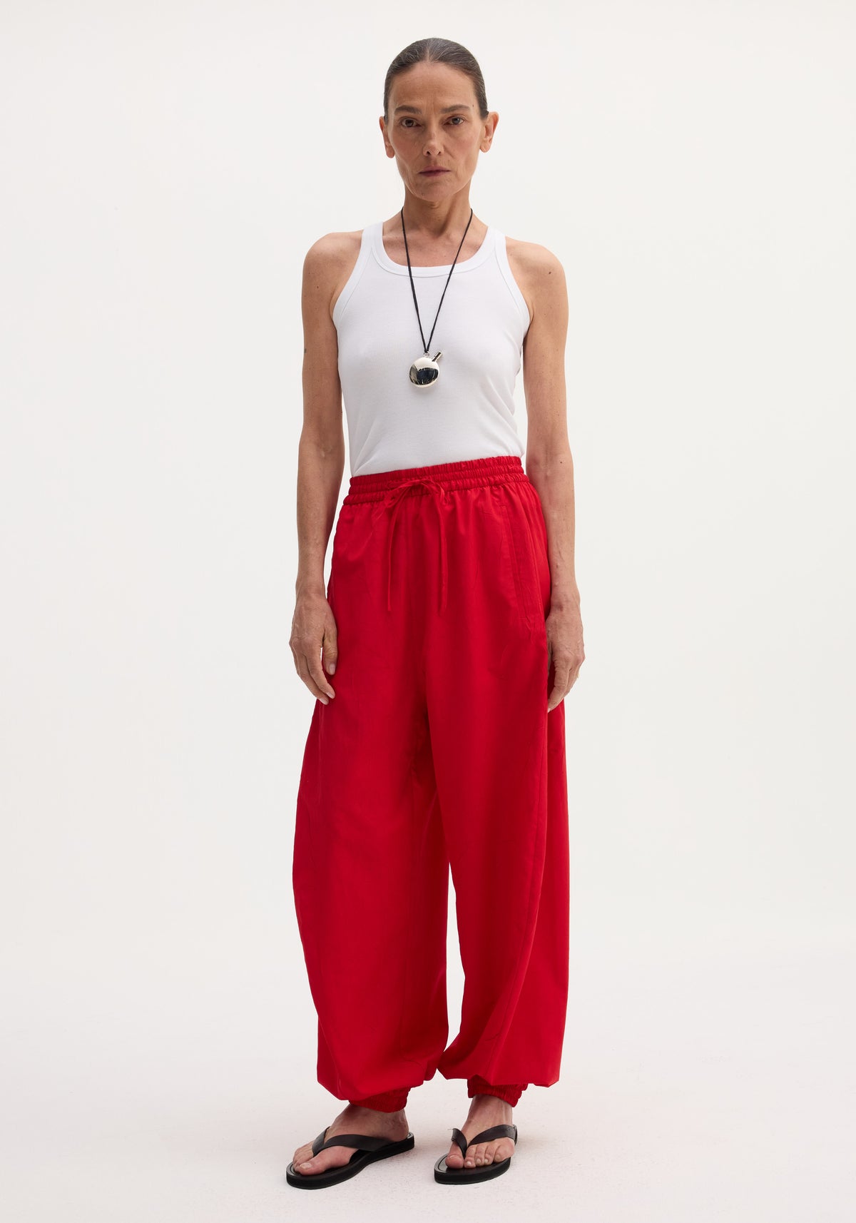 Woman wearing red Balloon track pants | red with elastic waistband and drawstring, paired with a white sleeveless top and black sandals, standing against a plain background.