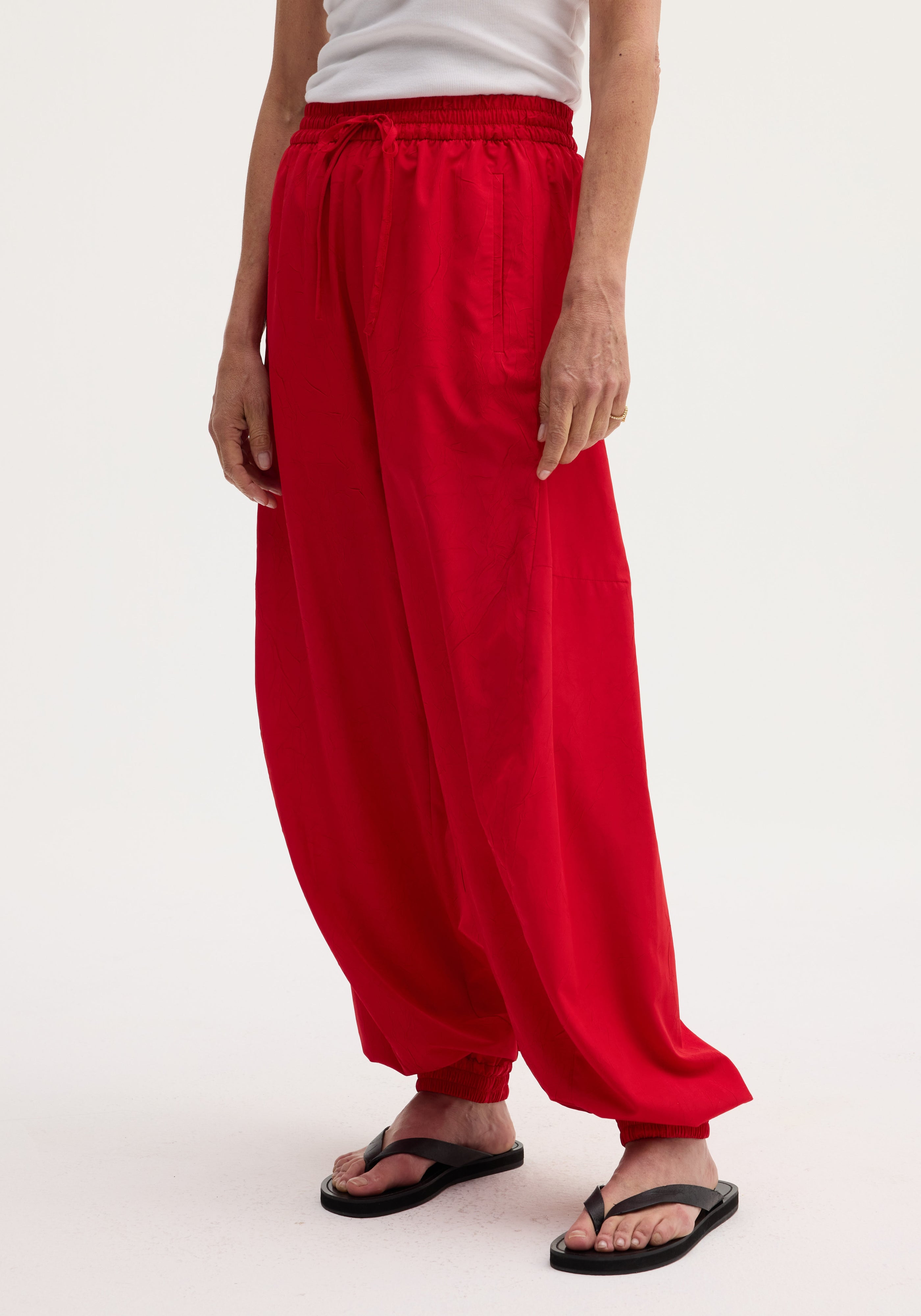Side view of a person wearing bright red Balloon track pants | red with a loose, puffy fit around the legs, gathered elastic waistband, and elastic cuffs at the ankles paired with black flip-flops.
