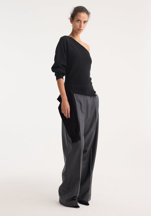 Model in an off-shoulder single sleeve top | black, showcasing its asymmetric design with a draped hemline, paired with high-waisted, wide-leg gray trousers.
