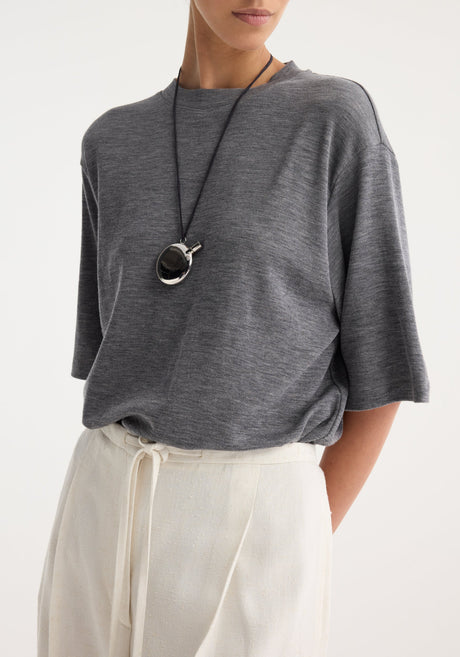 Woman wearing a heather-gray tee and off-white trousers, featuring a silver perfume bottle necklace with a shiny two-tone pendant on a black cord.