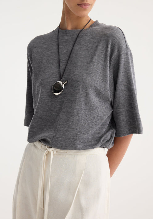 Woman wearing a heather-gray tee and off-white trousers, featuring a silver perfume bottle necklace with a shiny two-tone pendant on a black cord.