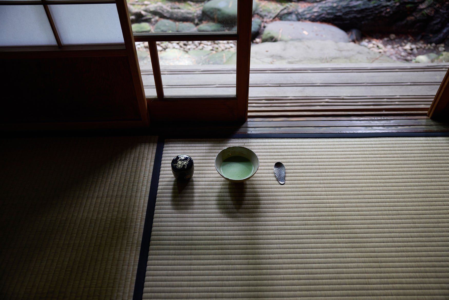 Bowl of matcha with a hammered spoon next to it on a tatami mat in the window of a Japanese tea house.