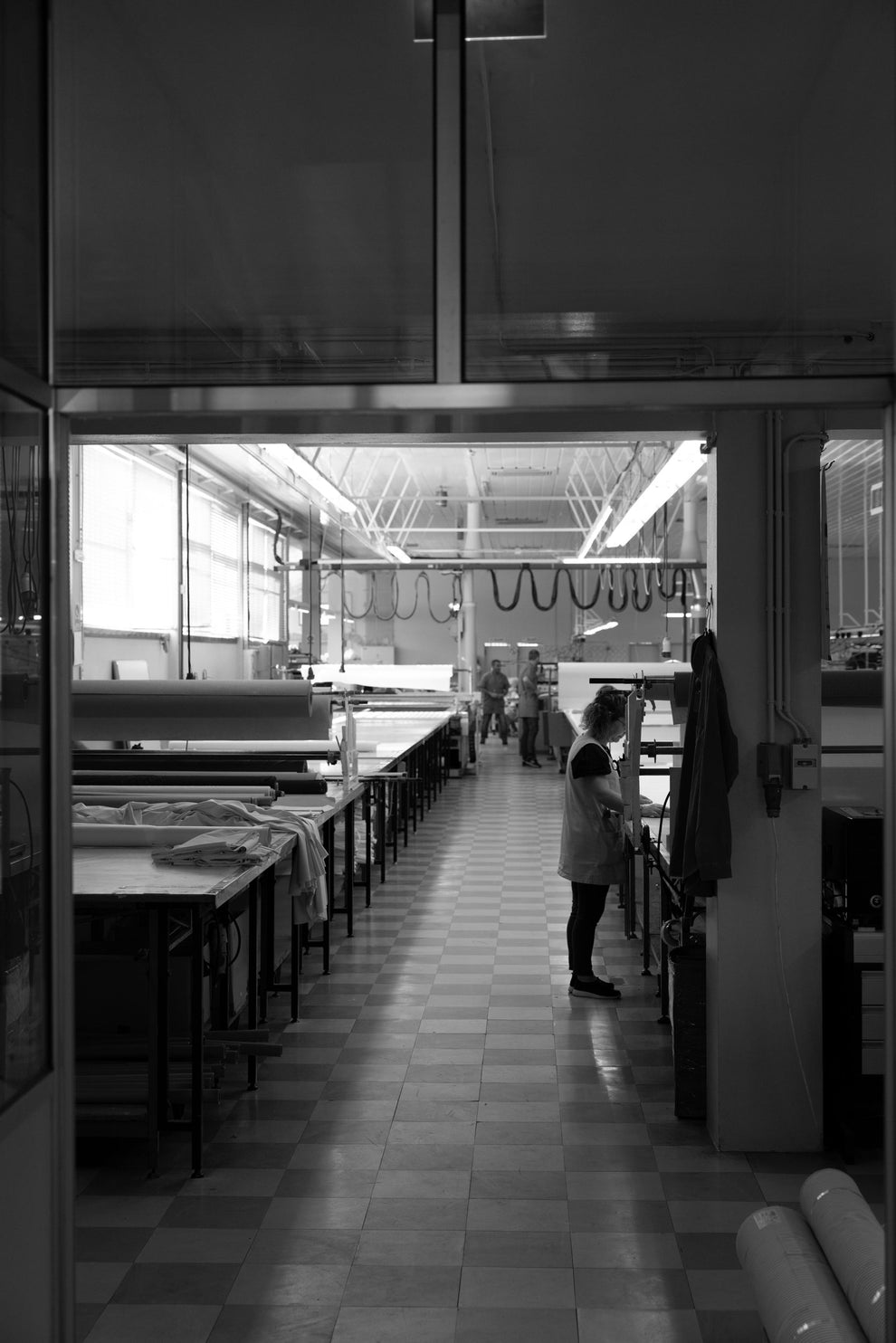 Long industrial space part of a garment factory with workers and large equipment and a tiled floor.