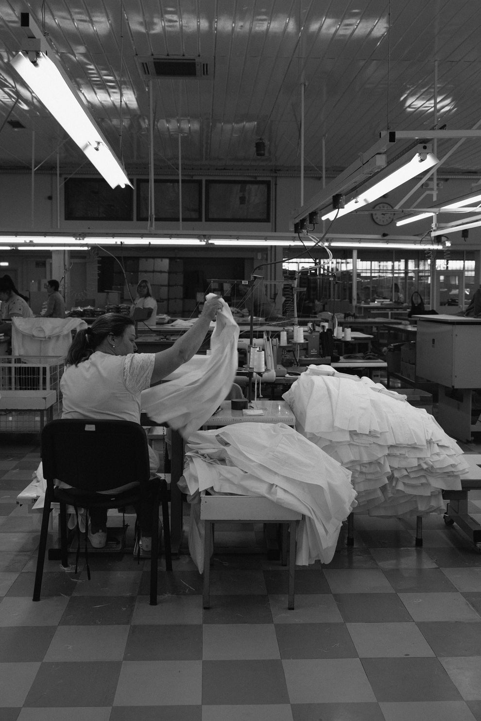 Factory interior with woman behind a sewing machine sewing shirts and stacking them next to her.
