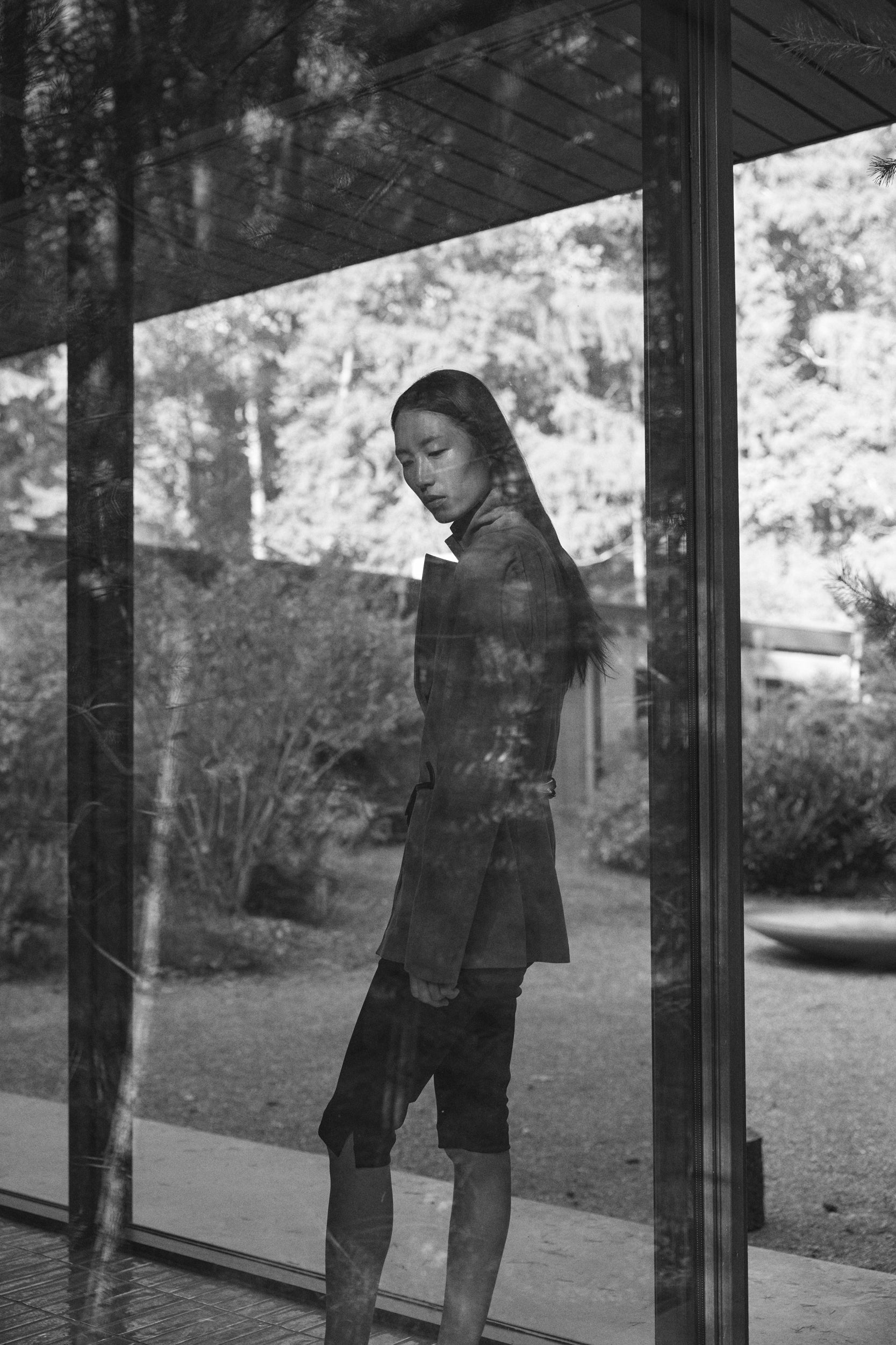 Person standing in a reflective glass structure with a garden view wearing a suede blazer and capri leggings