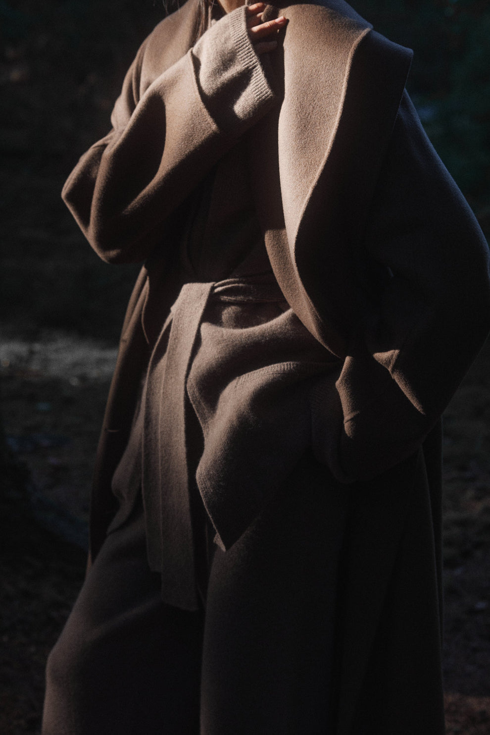 Person wearing a taupe coat layered over a taupe knitted belted cardigan against a dark background