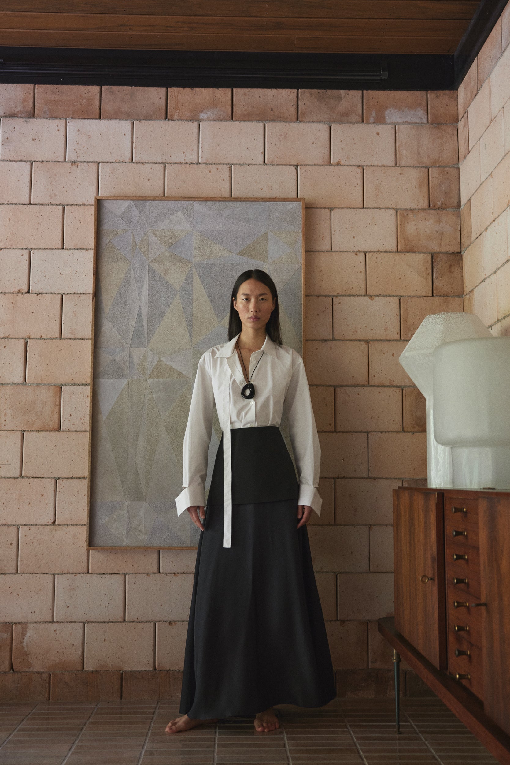 Woman in a white blouse and black long silk skirt standing in a room with a brick wall and abstract painting.