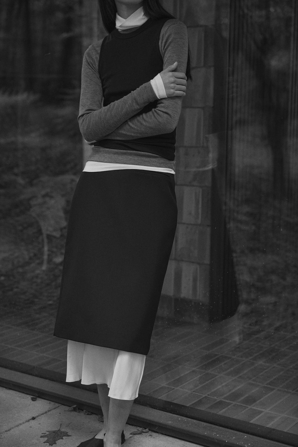 Woman wearing layered merino wool tops and a black skirt leaning against a window with forest setting reflected in it
