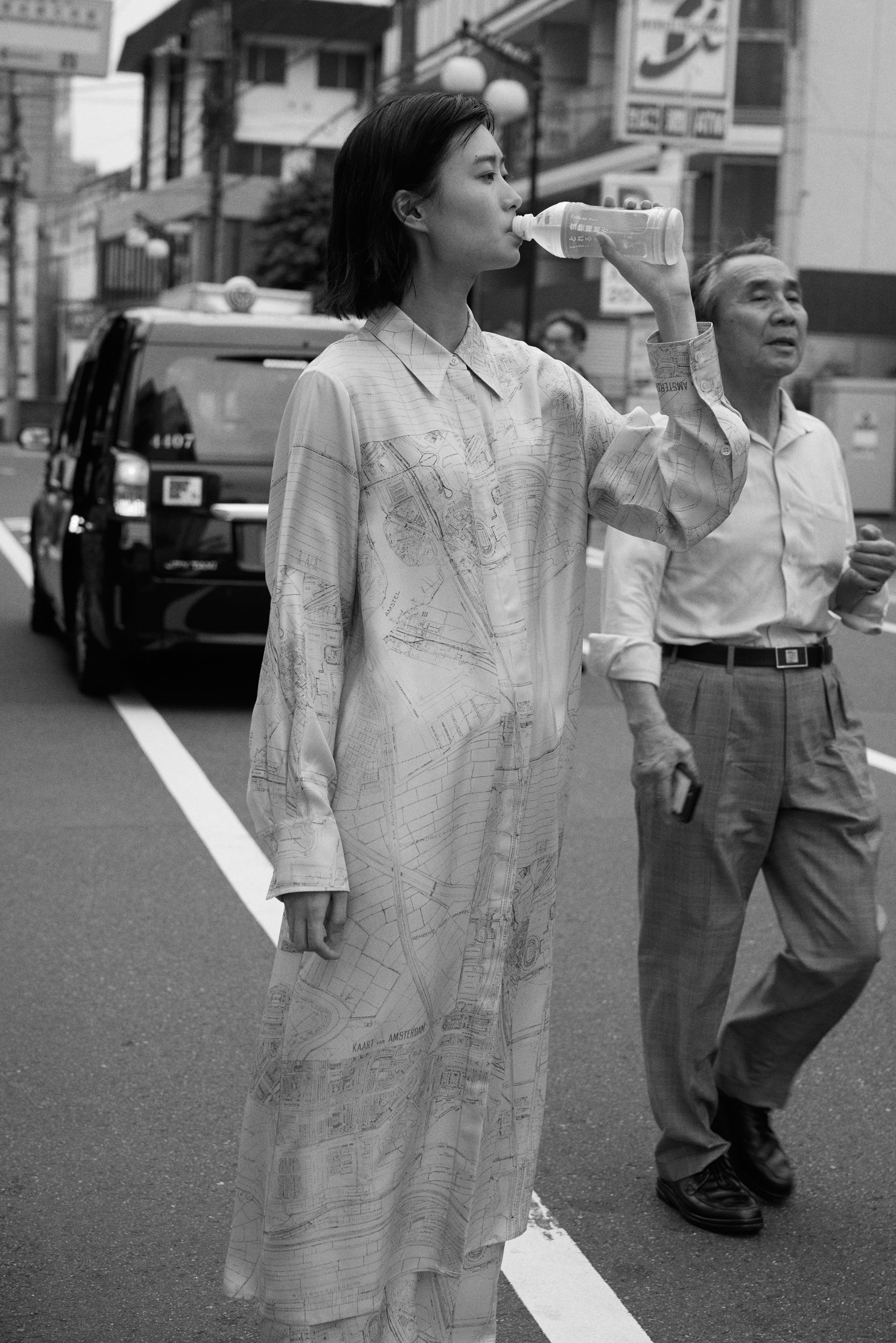 Woman standing in a busy street in a dress with a print of a drawn map, drinking a bottle of water. 