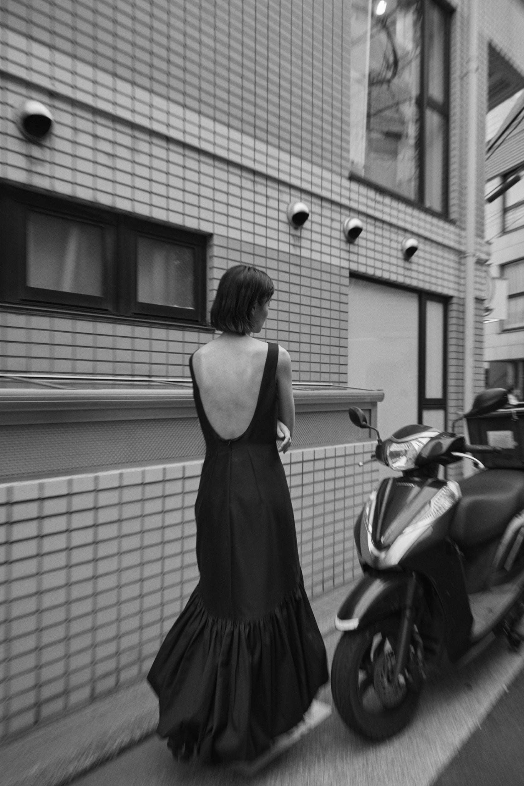 Woman photographed from the back in a small Tokyo alley wearing a black voluminous evening dress. 