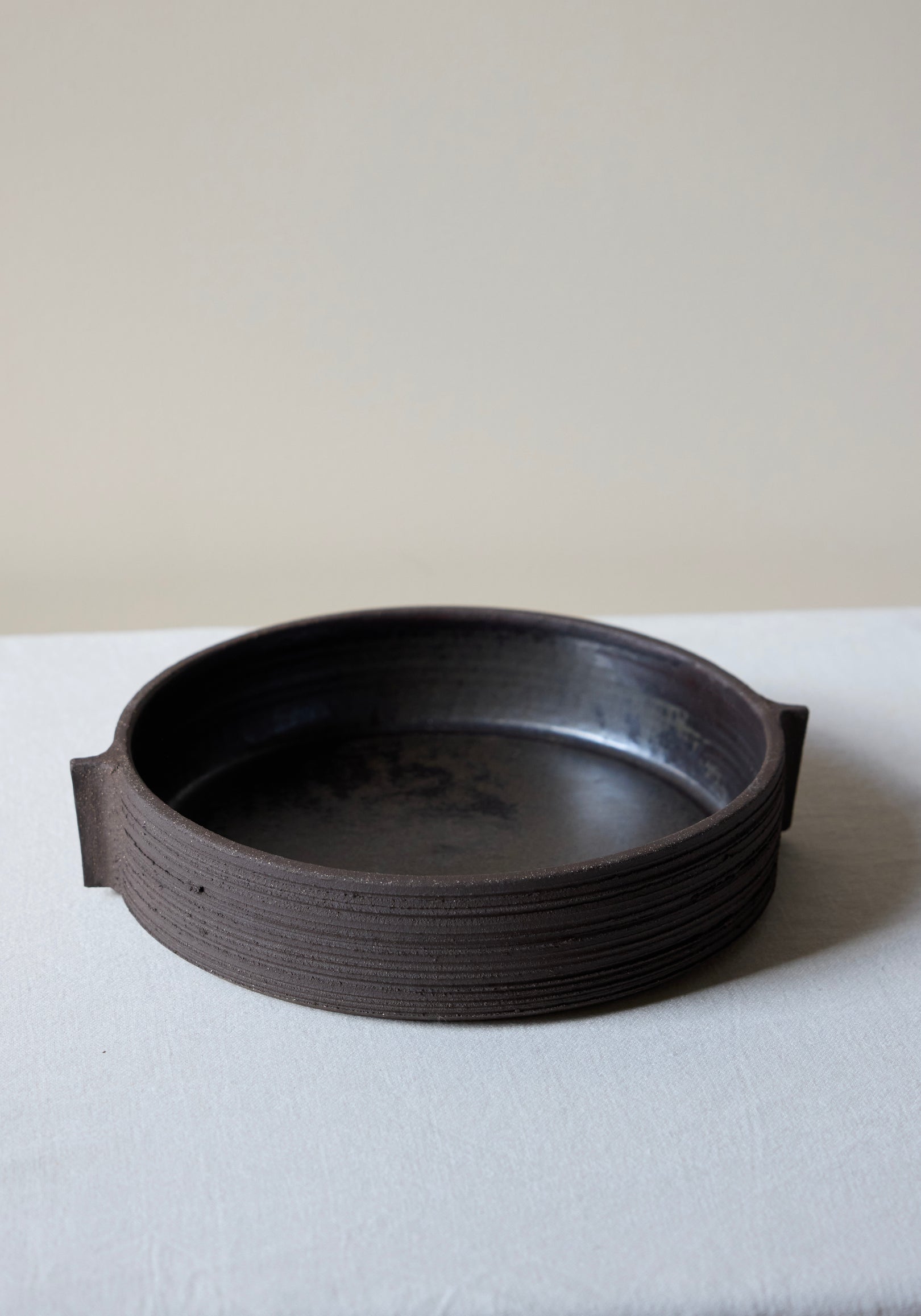 Round dark brown textured ceramic serving plate with carved sides and small handle-like extensions on each side, shown on a light fabric surface, Carved Ceramic Serving Plate | soil.