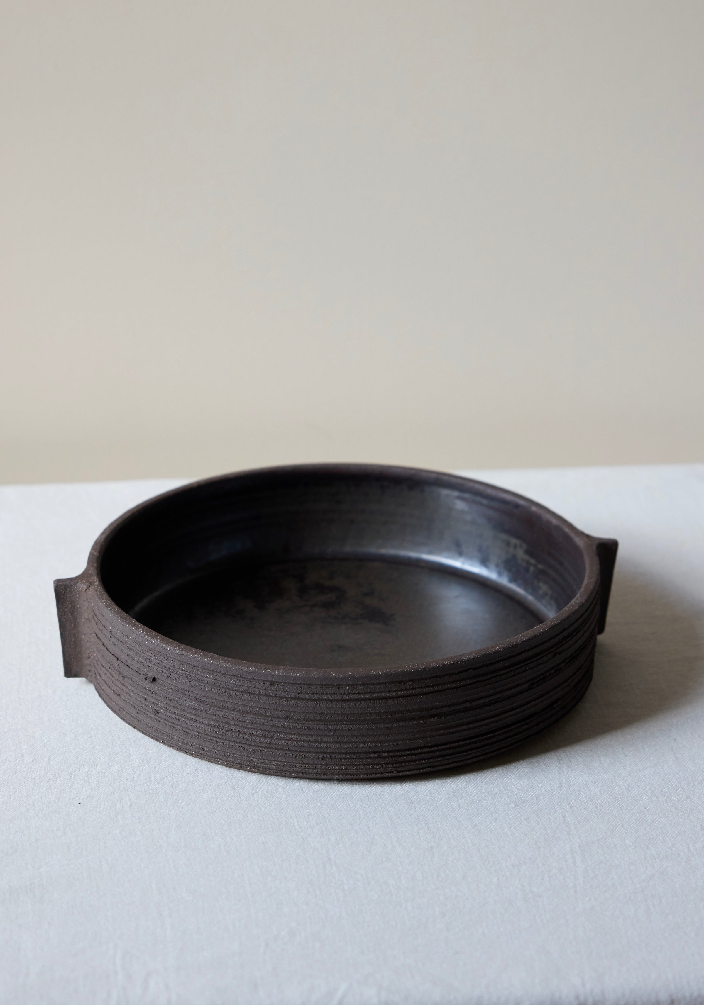 Round dark brown textured ceramic serving plate with carved sides and small handle-like extensions on each side, shown on a light fabric surface, Carved Ceramic Serving Plate | soil.