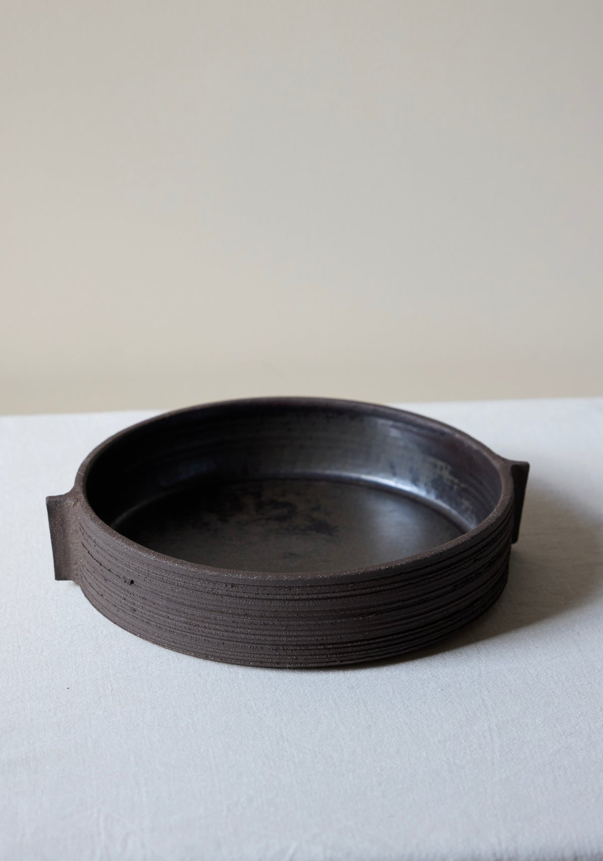Round dark brown textured ceramic serving plate with carved sides and small handle-like extensions on each side, shown on a light fabric surface, Carved Ceramic Serving Plate | soil.
