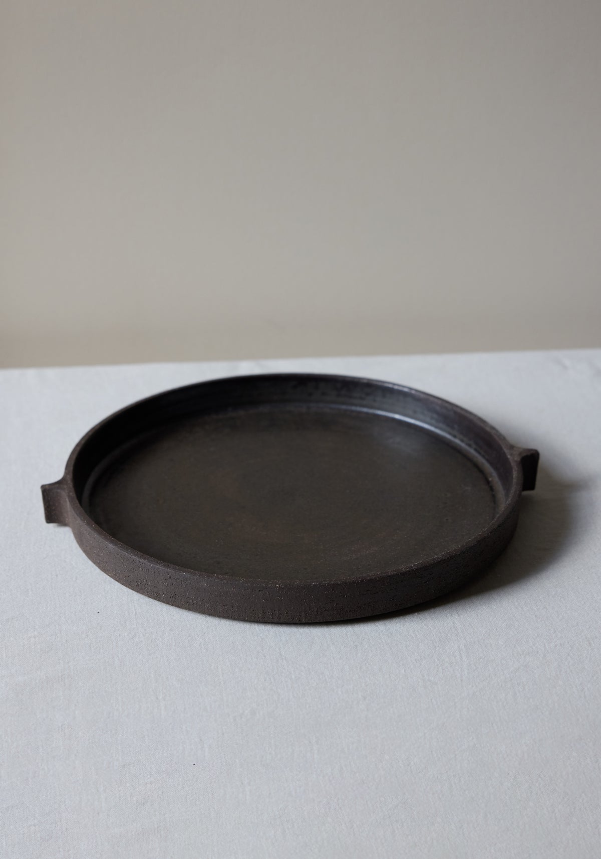 Ceramic Serving Plate | soil in a matte dark brown finish with a minimalist round design and two small handles, placed on a light-colored fabric surface.