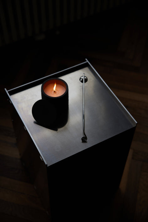 Candle Stopper | silver: A lit candle on a steel table accompanied by a spiraling stainless steel candle snuffer, elegantly designed for safe extinguishing.