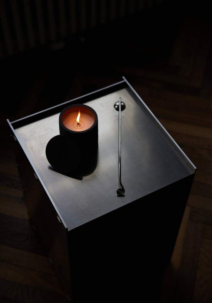 Candle Stopper | silver: A lit candle on a steel table accompanied by a spiraling stainless steel candle snuffer, elegantly designed for safe extinguishing.