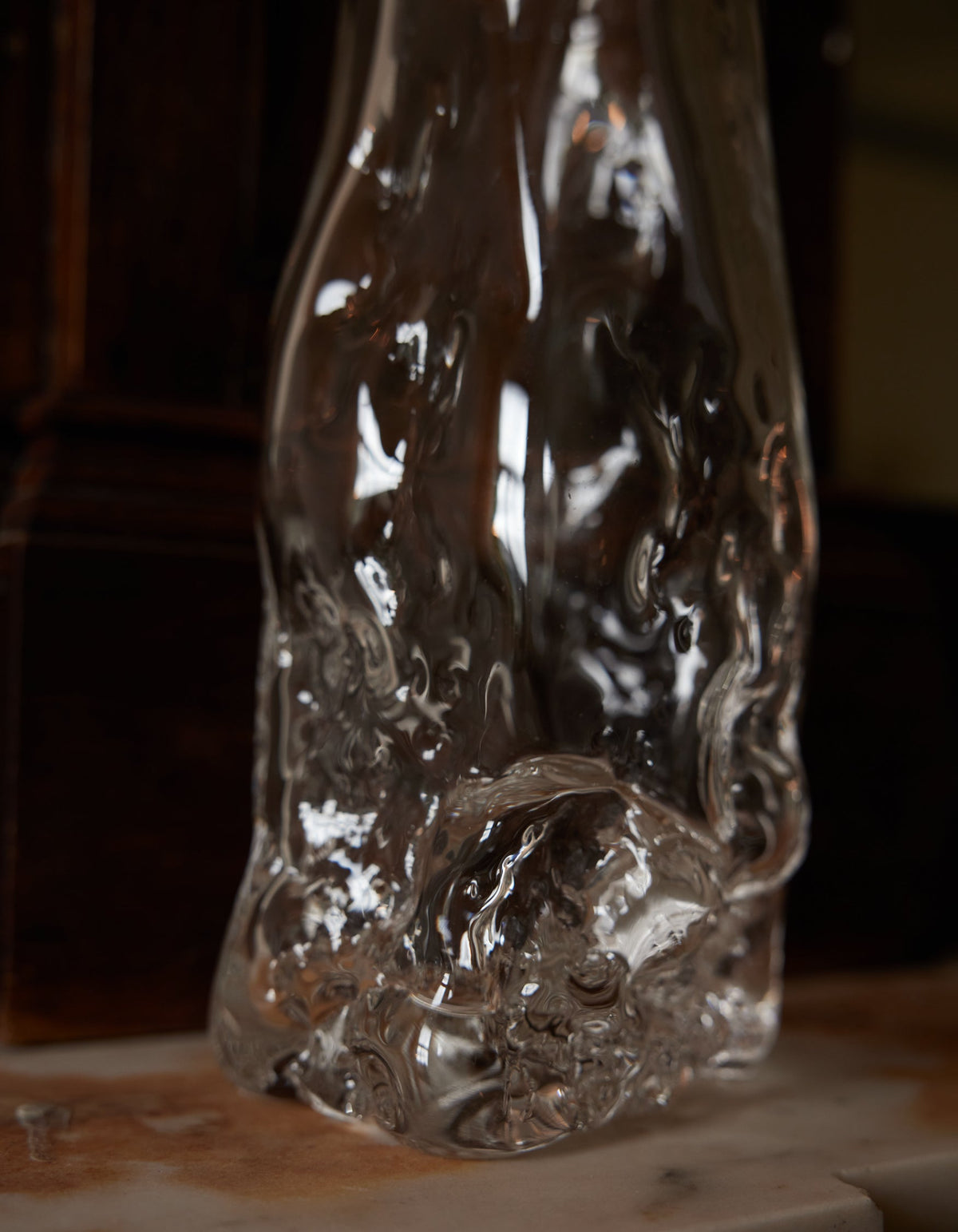 Blown vase | Glass: A clear, irregularly shaped glass vase with fluid, rippled design, showcasing intricate craftsmanship, sitting on a smooth surface against a wooden backdrop.