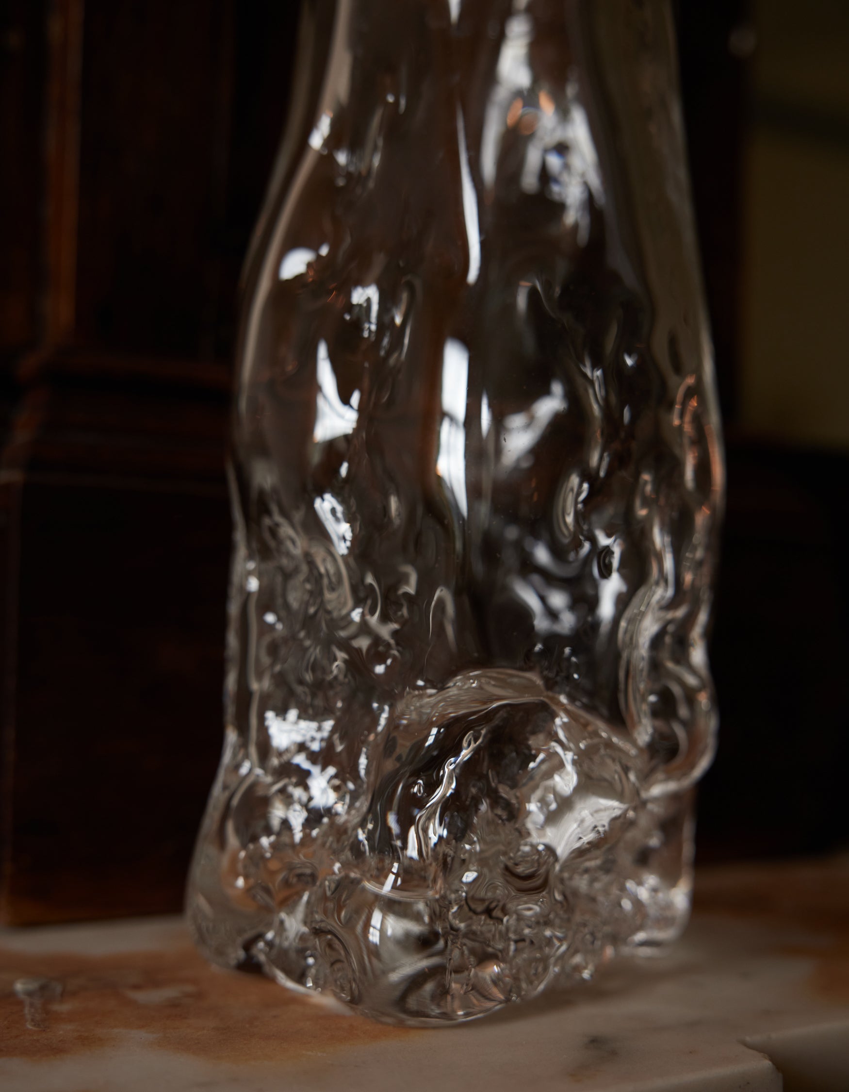Blown vase | Glass: A clear, irregularly shaped glass vase with fluid, rippled design, showcasing intricate craftsmanship, sitting on a smooth surface against a wooden backdrop.