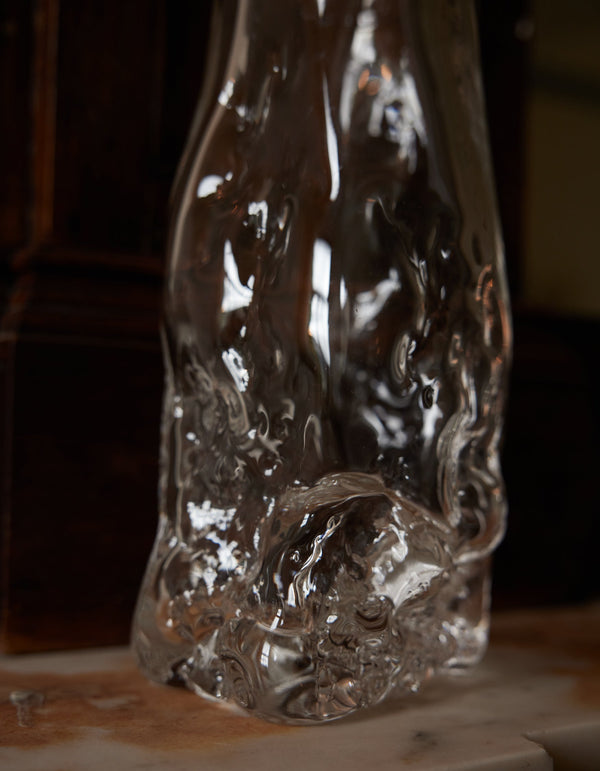 Blown vase | Glass: A clear, irregularly shaped glass vase with fluid, rippled design, showcasing intricate craftsmanship, sitting on a smooth surface against a wooden backdrop.