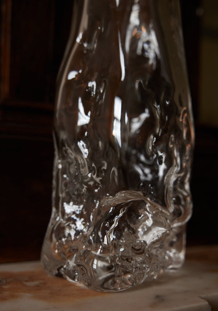 Blown vase | Glass: A clear, irregularly shaped glass vase with fluid, rippled design, showcasing intricate craftsmanship, sitting on a smooth surface against a wooden backdrop.