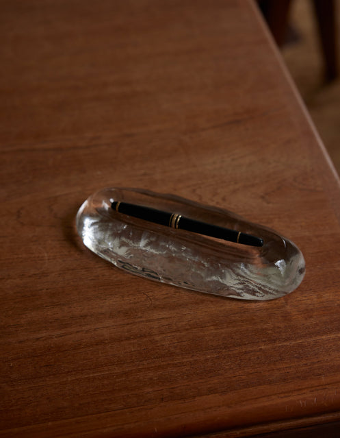 Close-up of Long trinket | Glass, an elegant clear glass pen rest, featuring a black pen with gold trim on a wooden desk.