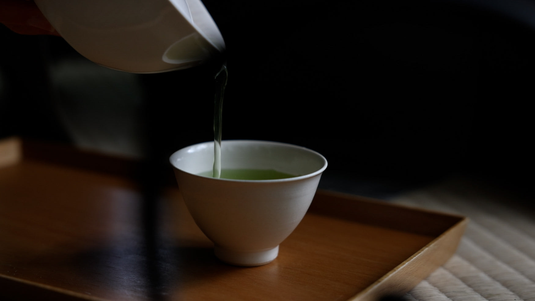 Close up of green tea being poured into a small white porcelain cup placed on top of a wooden tray. 