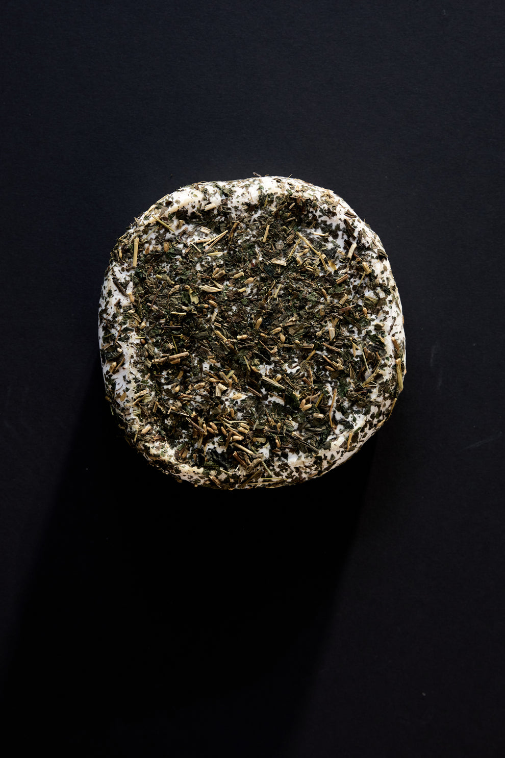 Image of a round cheese covered in dried herbs on a black background