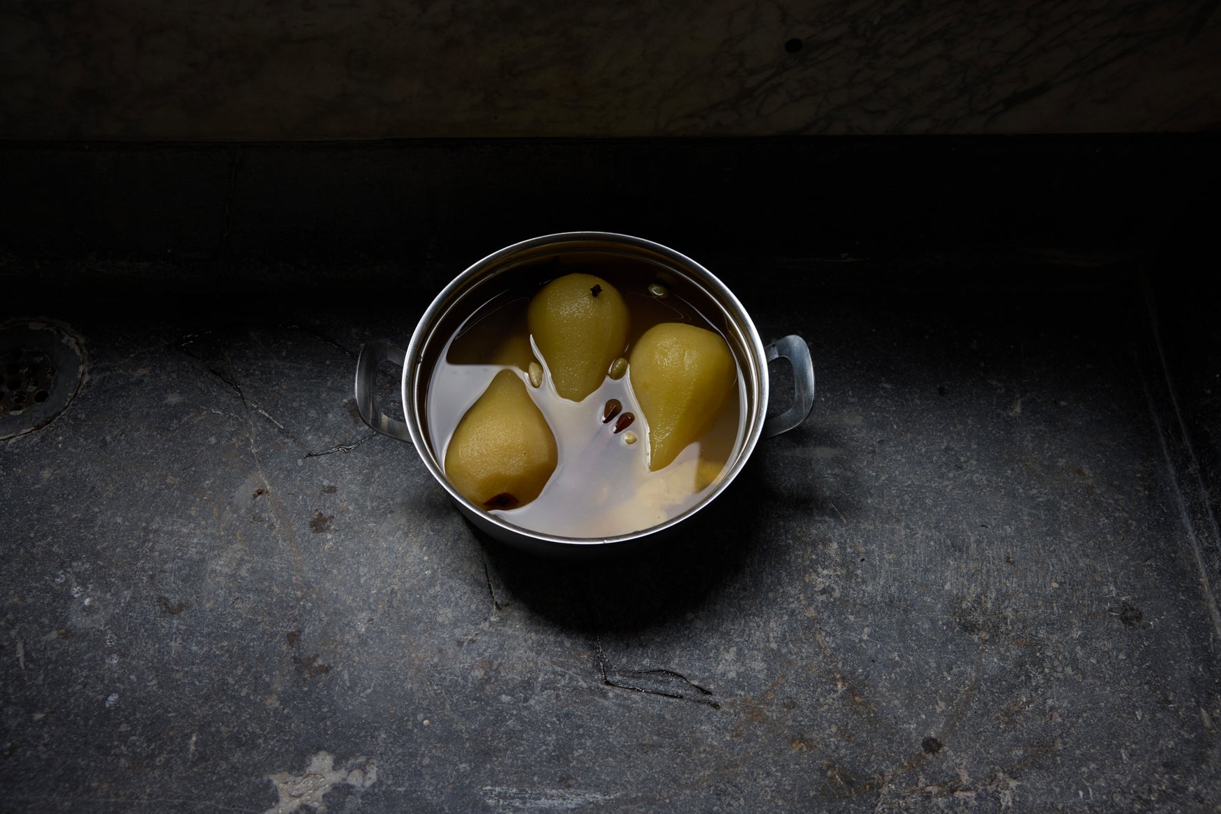 Three pears submerged in a pan of liquid on a dark grey stone surface 