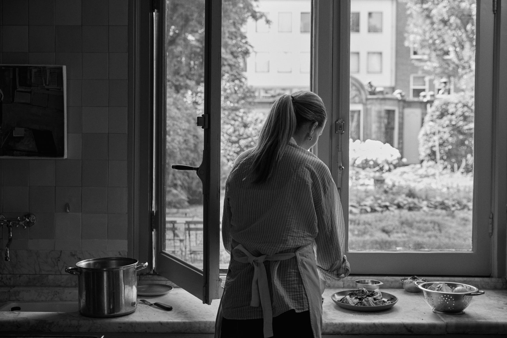 Person standing at a marble countertop with pans and plates on it, in front of a window cooking a dish 