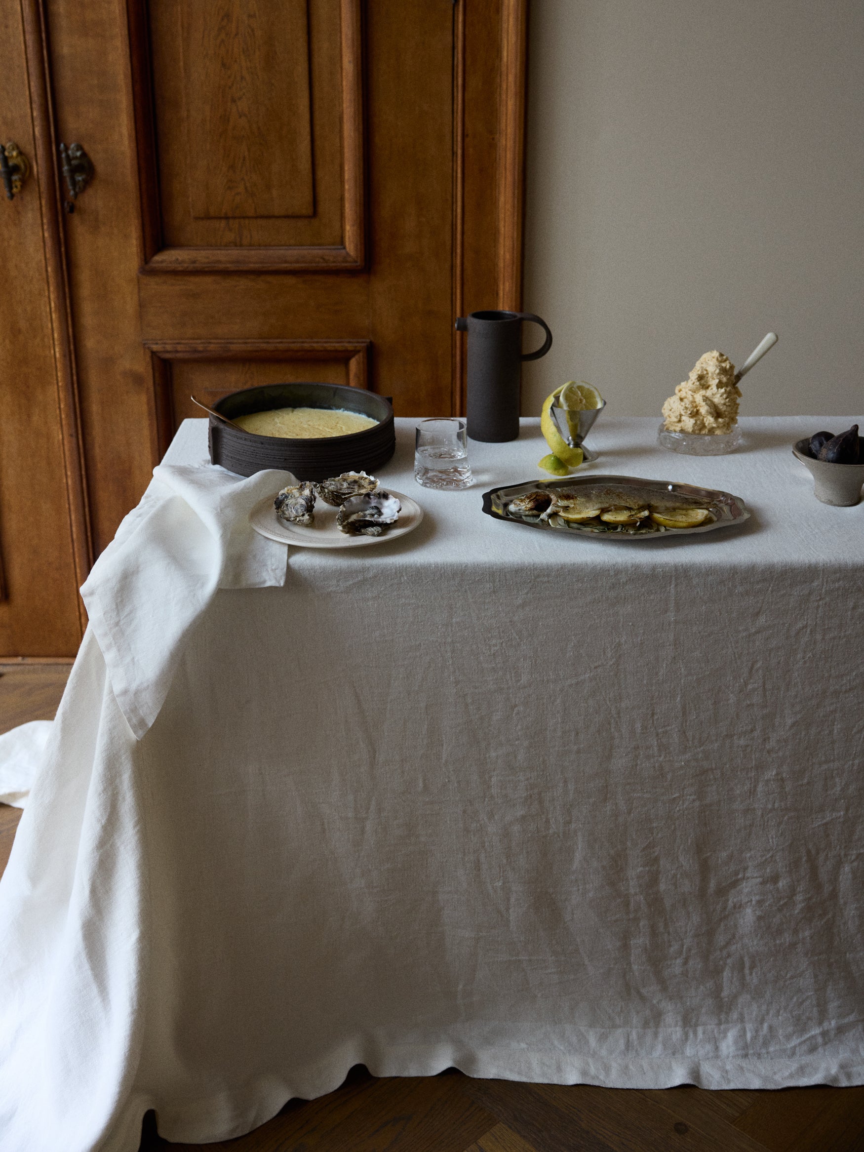 Corner of a table with linnen tablecloth with oysters, a bowl of custard and a carafe on it against a wooden backdrop