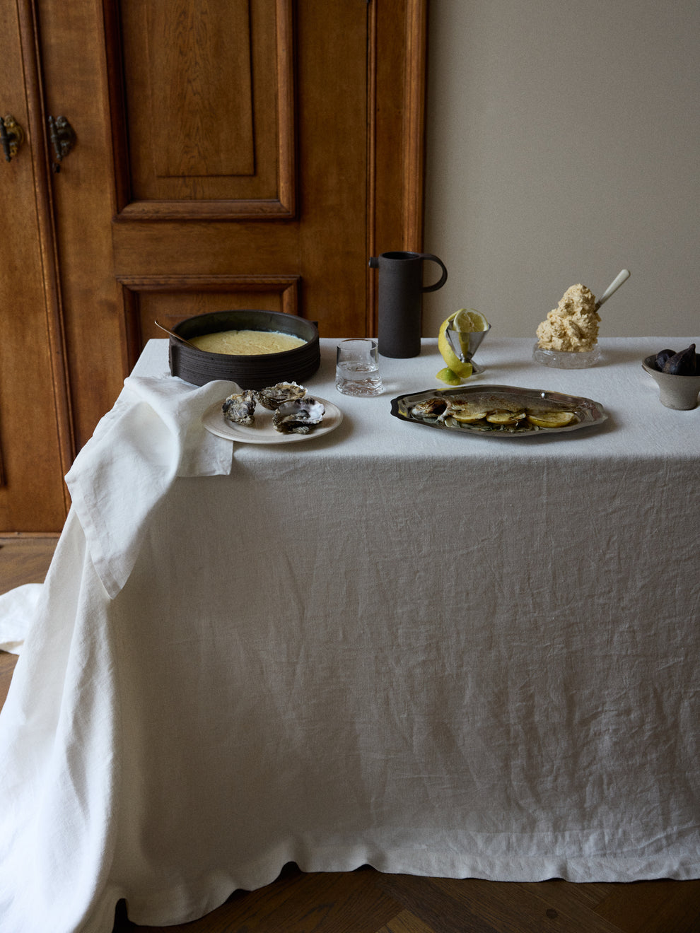 Corner of a table with linnen tablecloth with oysters, a bowl of custard and a carafe on it against a wooden backdrop