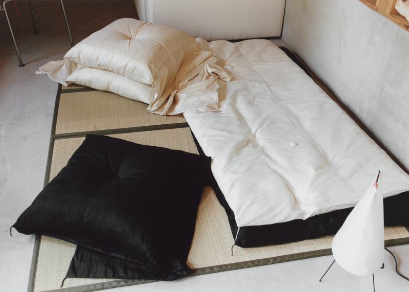 A minimalist Japanese-style tatami floor mattress setup with a white quilted mattress topper, cream bedding, a black floor cushion, and a woven tatami mat on a light floor.