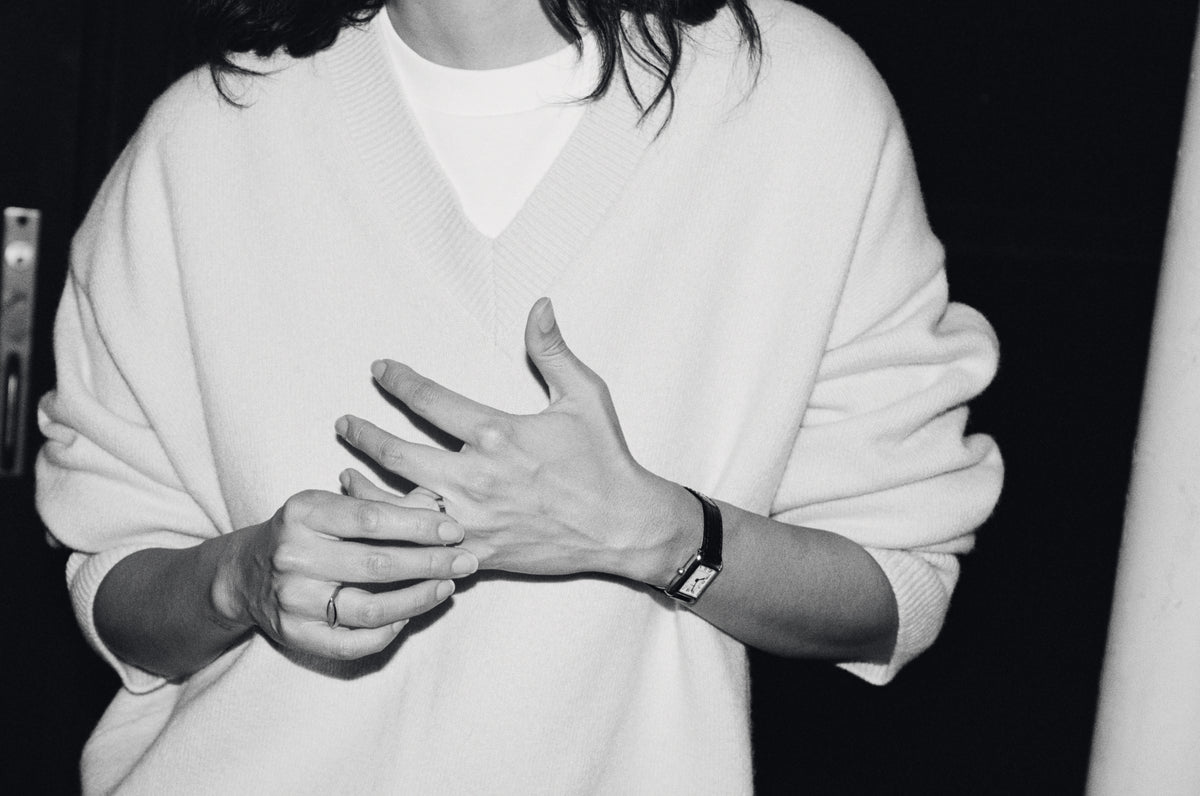 A black and white close-up of a person wearing a white knit V-neck sweater over a white top, with hands clasped in front and a watch on one wrist.