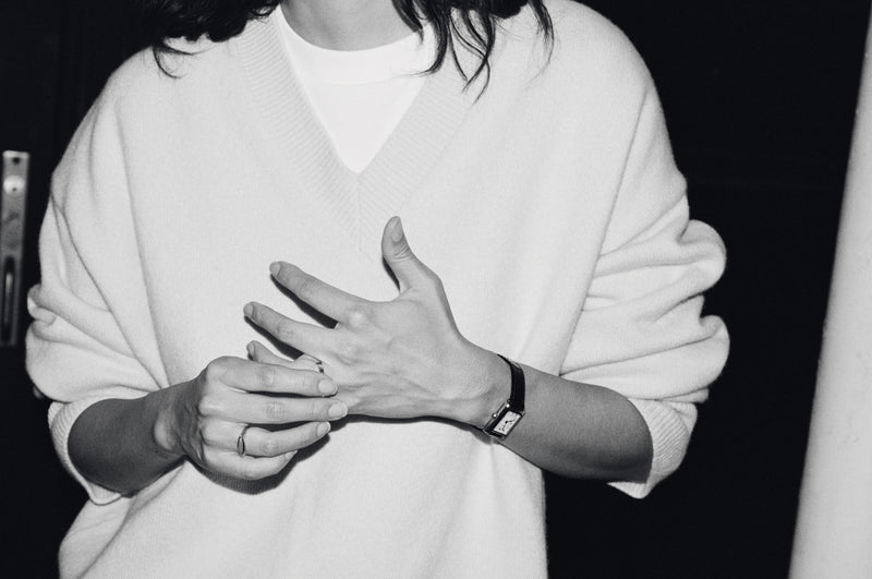 A black and white close-up of a person wearing a white knit V-neck sweater over a white top, with hands clasped in front and a watch on one wrist.