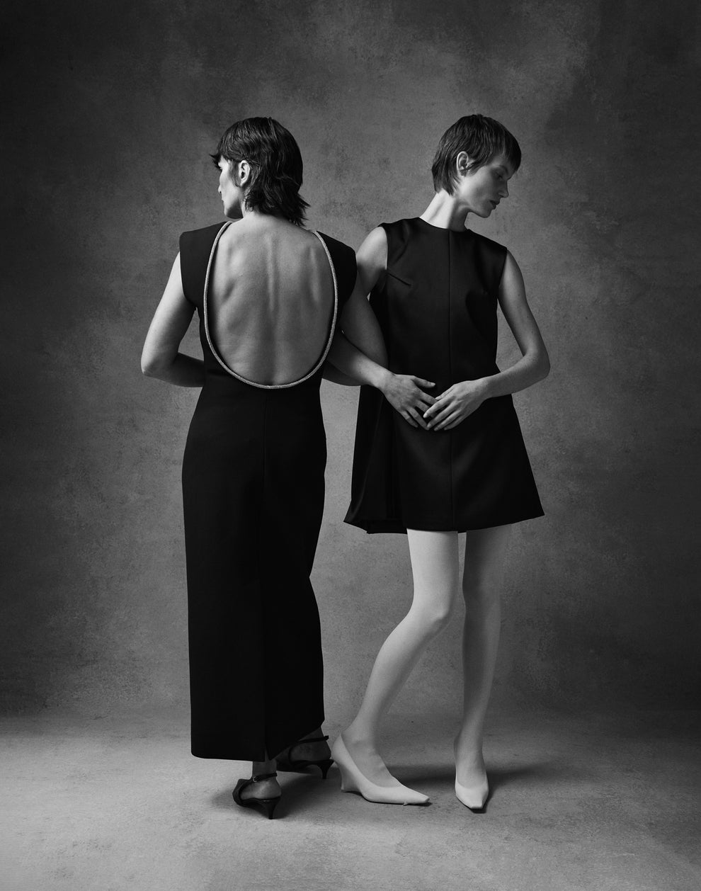 Two woman with hooked arms, one from the back in a long black low back dress, one from the front in a short a-line black dress