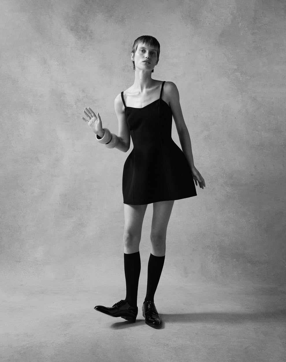 Woman wearing a short black spaghetti strap dress in a sculptural shape with knee high black socks and lacquered shoes